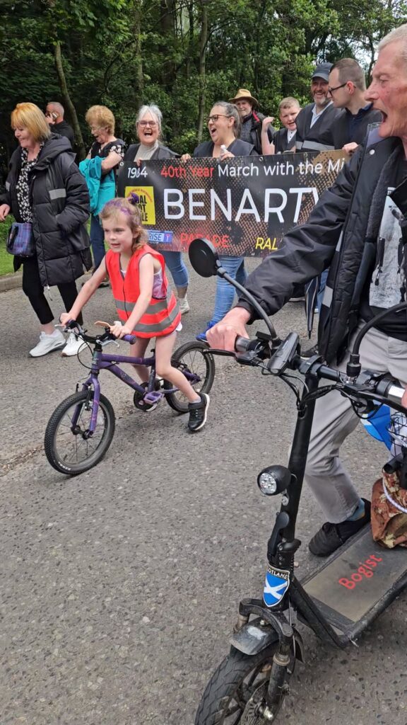 A protest that has become intergenerational: Maeve rides her bike beside Sean Lee on his mobility scooter and joins in with her grandfather’s words, ‘Maggie, Maggie, Maggie, Out! Out! Out!’, Benarty, 15 June 2024.